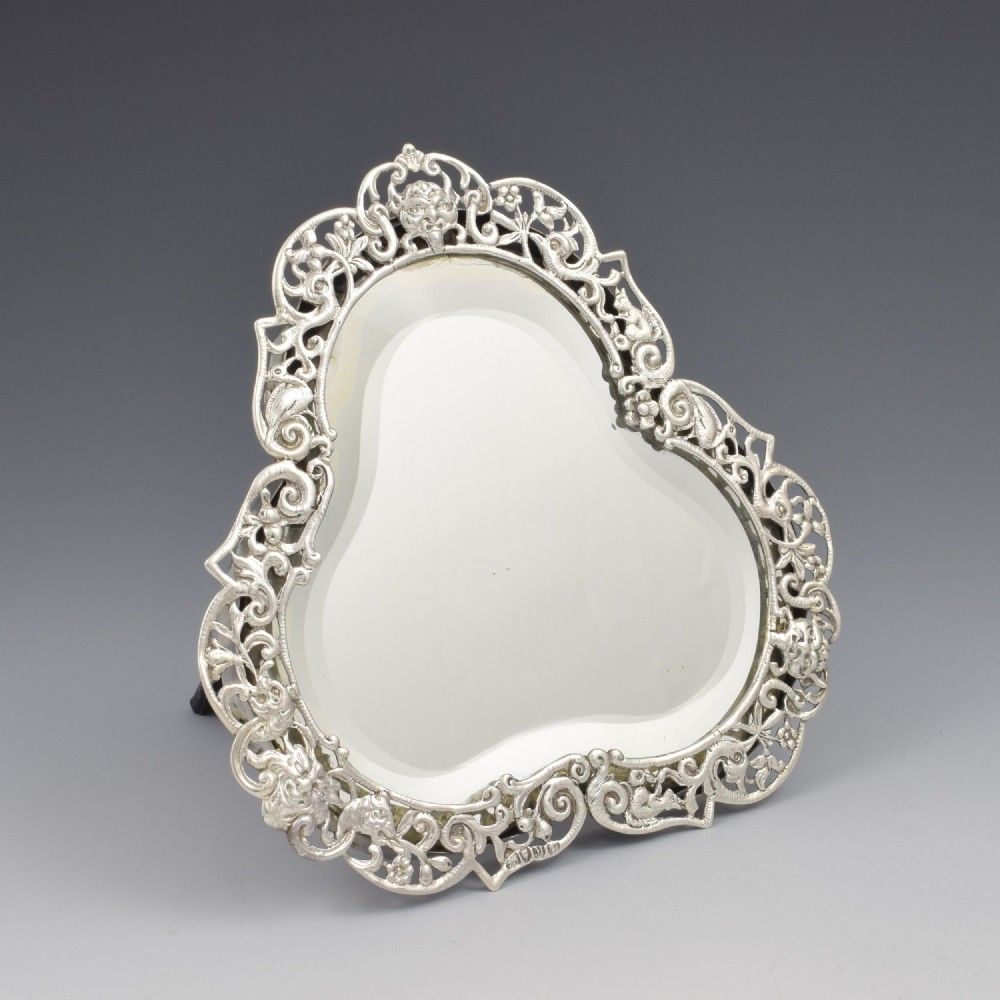 An antique silver dressing table mirror featuring intricate pierced scrollwork, foliage, bird motifs, and a mascaron crest around a shaped glass plate.