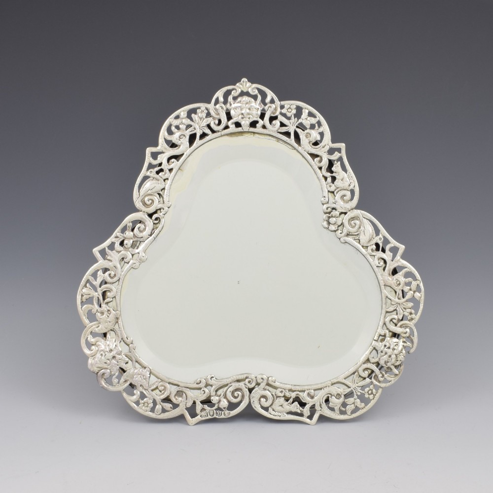 An antique silver dressing table mirror featuring intricate pierced scrollwork, foliage, bird motifs, and a mascaron crest around a shaped glass plate. View II