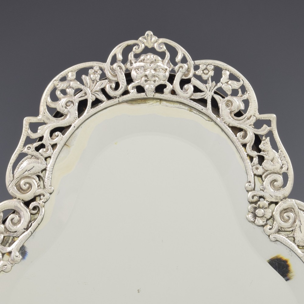 An antique silver dressing table mirror featuring intricate pierced scrollwork, foliage, bird motifs, and a mascaron crest around a shaped glass plate. View III
