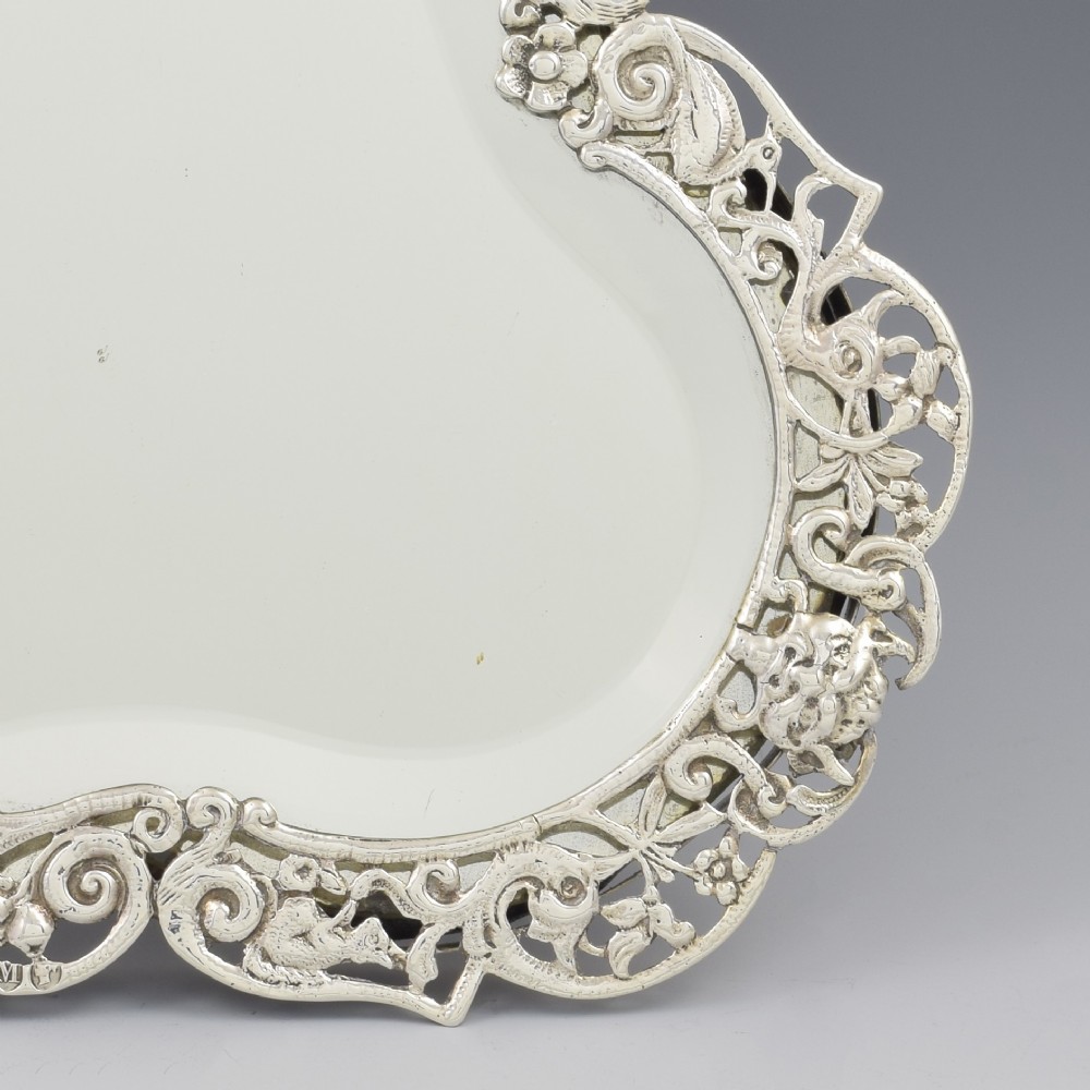 An antique silver dressing table mirror featuring intricate pierced scrollwork, foliage, bird motifs, and a mascaron crest around a shaped glass plate. View VI