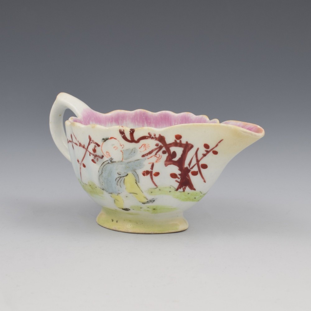 A small 18th-century soft-paste porcelain sauce boat featuring a scalloped fluted rim, loop handle, and hand-painted Chinoiserie figures with a pink interior border. View II