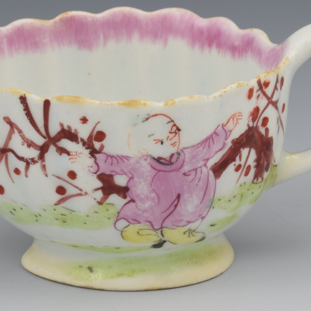 A small 18th-century soft-paste porcelain sauce boat featuring a scalloped fluted rim, loop handle, and hand-painted Chinoiserie figures with a pink interior border. View X