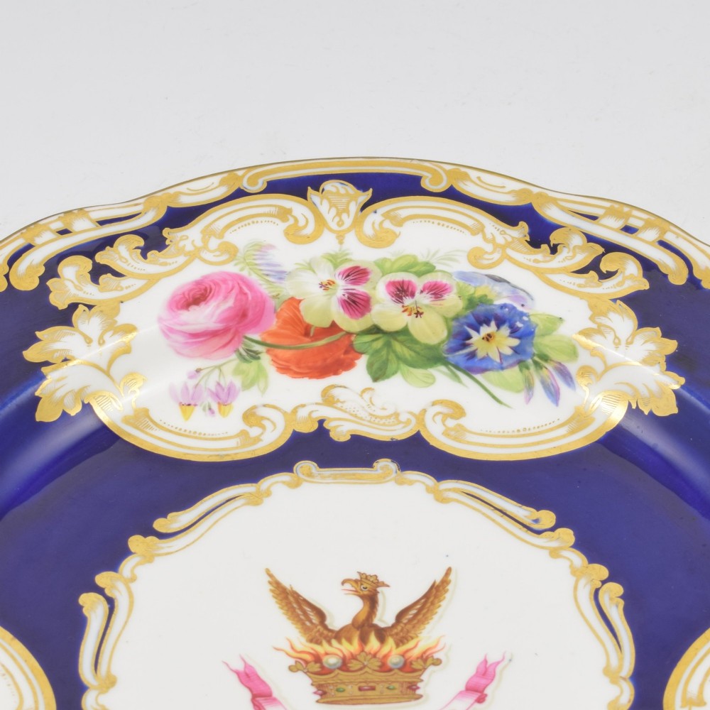 A 19th-century Copeland porcelain plate featuring a cobalt blue border with gilt scrollwork, hand-painted floral cartouches, and a central heraldic crest depicting a phoenix in flames above a coronet. View VII