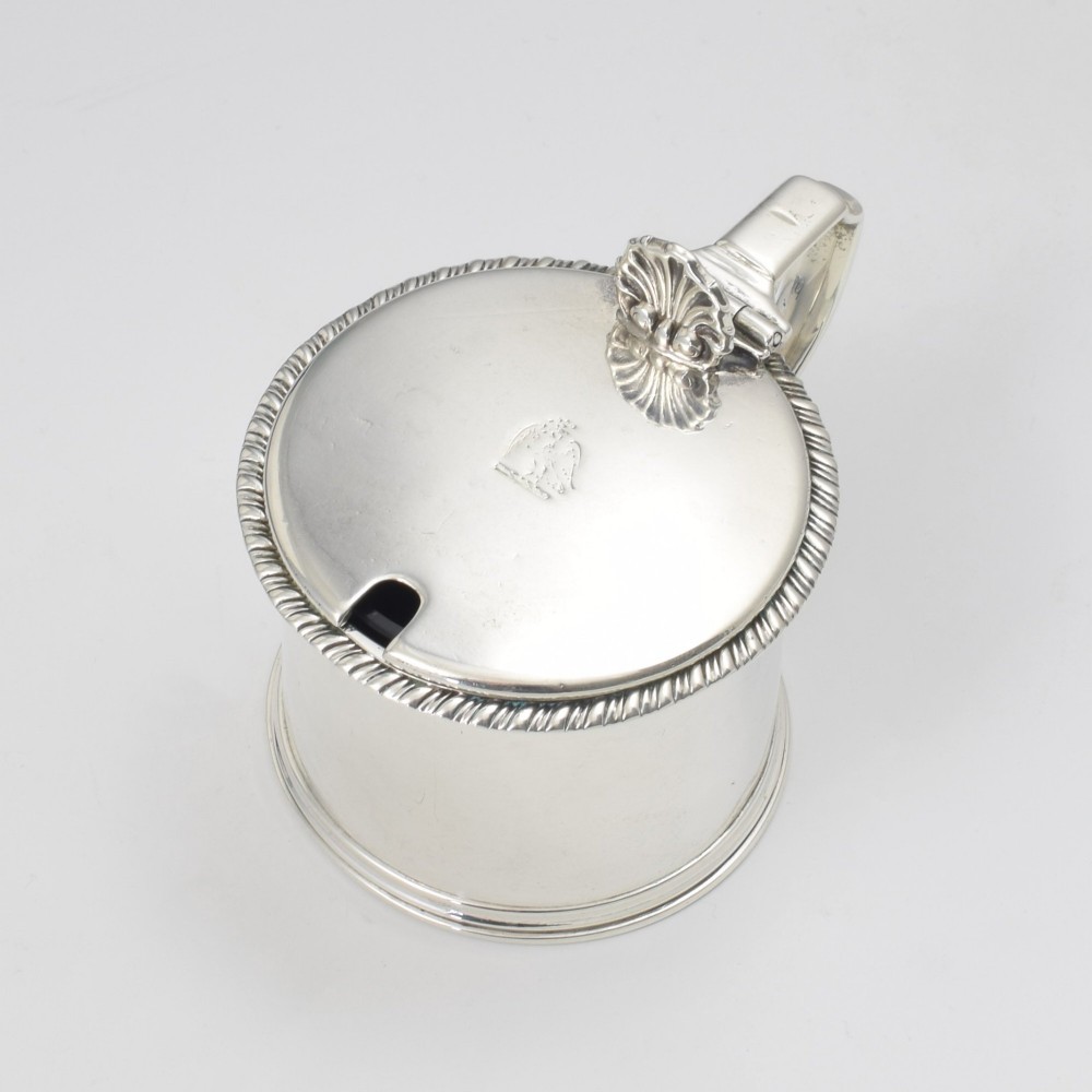 A cylindrical Georgian sterling silver drum mustard pot featuring a hinged lid, shell thumbpiece, and engraved crest.