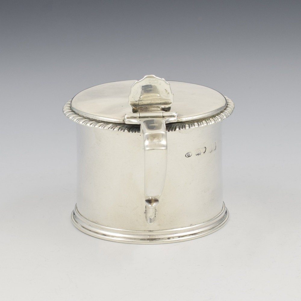 A cylindrical Georgian sterling silver drum mustard pot featuring a hinged lid, shell thumbpiece, and engraved crest. View X