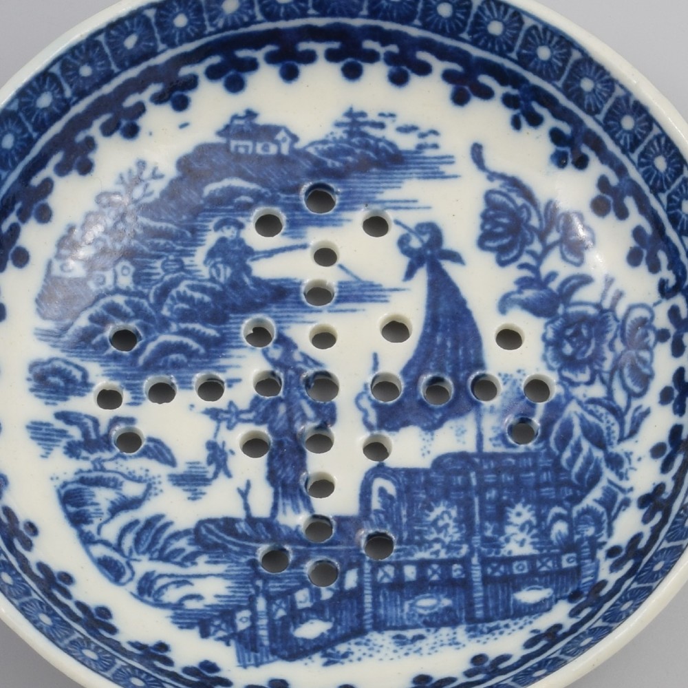 A circular blue and white transfer-printed porcelain egg drainer featuring a chinoiserie river scene with drainage perforations and a moulded shell handle. View II