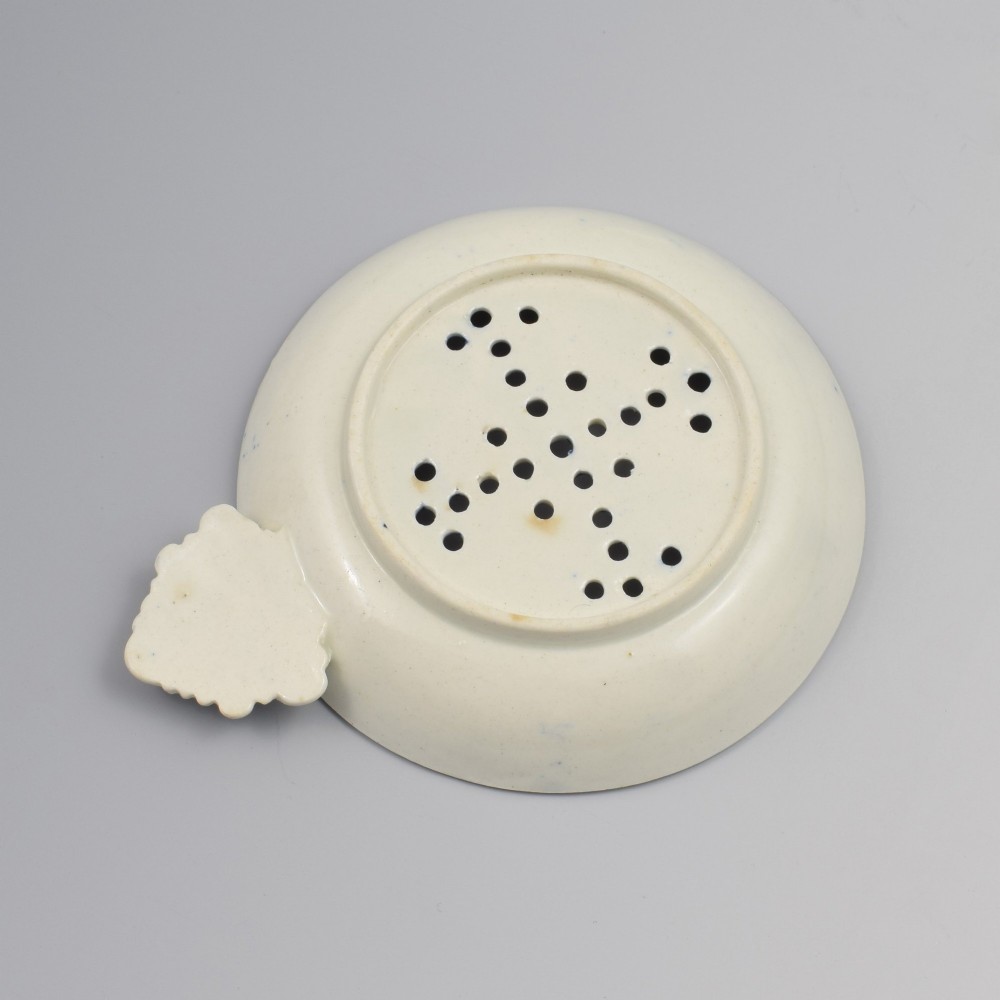 A circular blue and white transfer-printed porcelain egg drainer featuring a chinoiserie river scene with drainage perforations and a moulded shell handle. View III