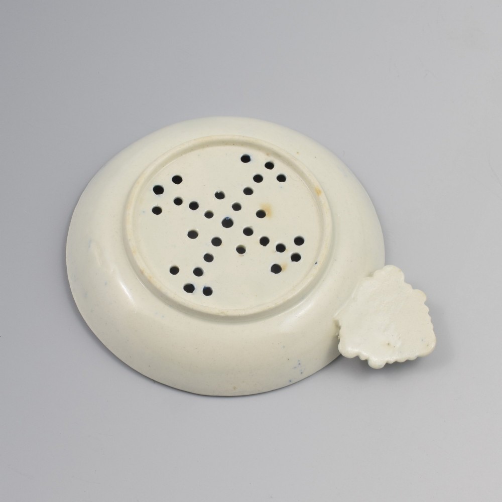 A circular blue and white transfer-printed porcelain egg drainer featuring a chinoiserie river scene with drainage perforations and a moulded shell handle. View IV