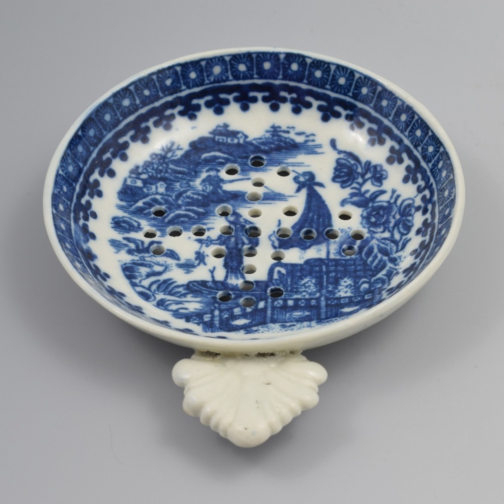 A circular blue and white transfer-printed porcelain egg drainer featuring a chinoiserie river scene with drainage perforations and a moulded shell handle. View V