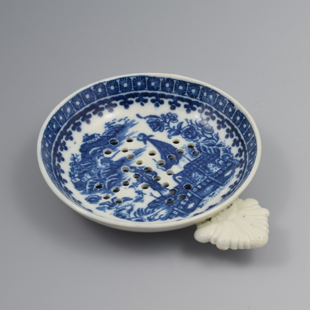 A circular blue and white transfer-printed porcelain egg drainer featuring a chinoiserie river scene with drainage perforations and a moulded shell handle. View VI