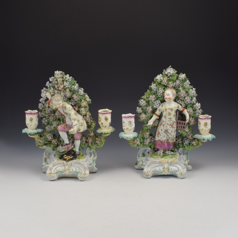 A pair of 18th-century Derby porcelain candlestick figures depicting children standing before flowering bocage trees set on scrollwork bases.