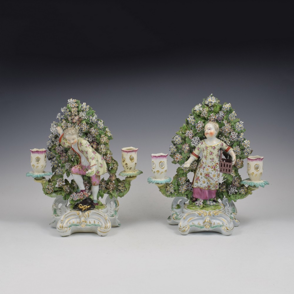 A pair of 18th-century Derby porcelain candlestick figures depicting children standing before flowering bocage trees set on scrollwork bases. View II