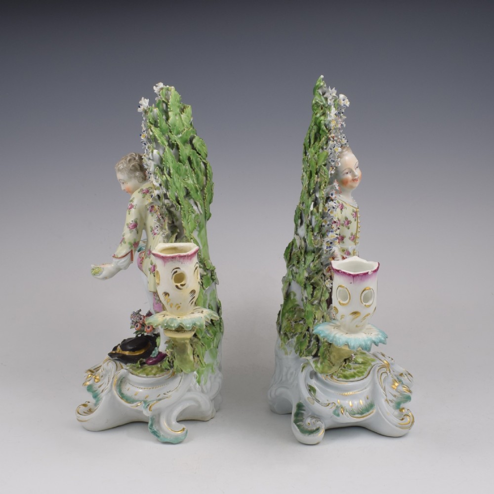 A pair of 18th-century Derby porcelain candlestick figures depicting children standing before flowering bocage trees set on scrollwork bases. View III