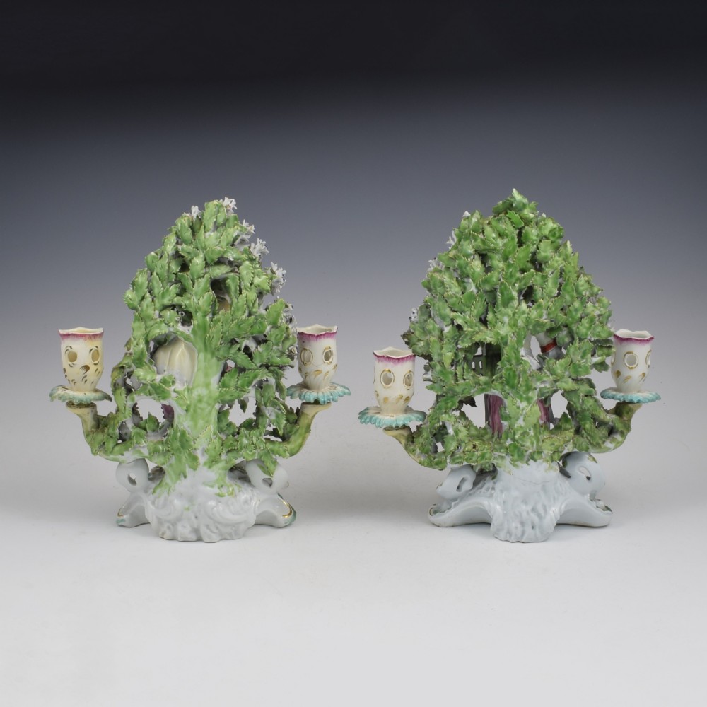 A pair of 18th-century Derby porcelain candlestick figures depicting children standing before flowering bocage trees set on scrollwork bases. View IV