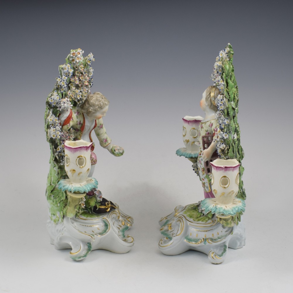 A pair of 18th-century Derby porcelain candlestick figures depicting children standing before flowering bocage trees set on scrollwork bases. View V