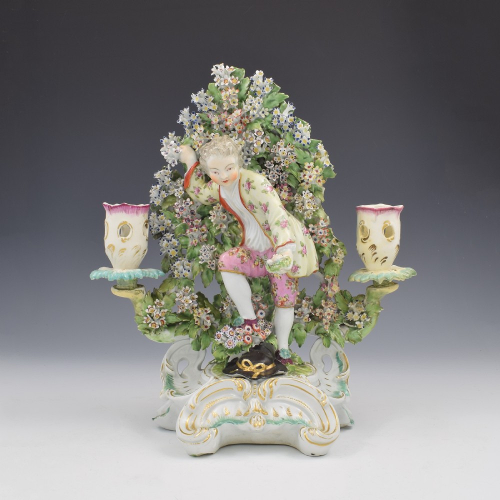 A pair of 18th-century Derby porcelain candlestick figures depicting children standing before flowering bocage trees set on scrollwork bases. View VI