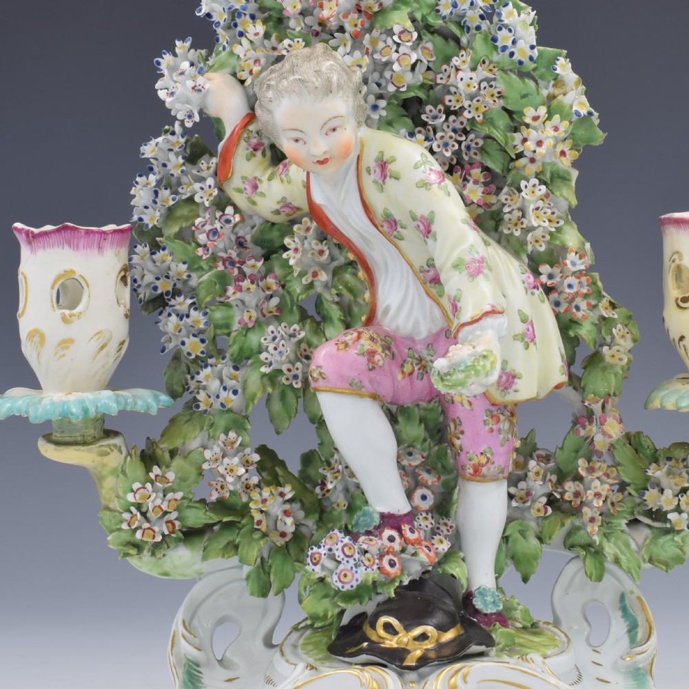A pair of 18th-century Derby porcelain candlestick figures depicting children standing before flowering bocage trees set on scrollwork bases. View VII
