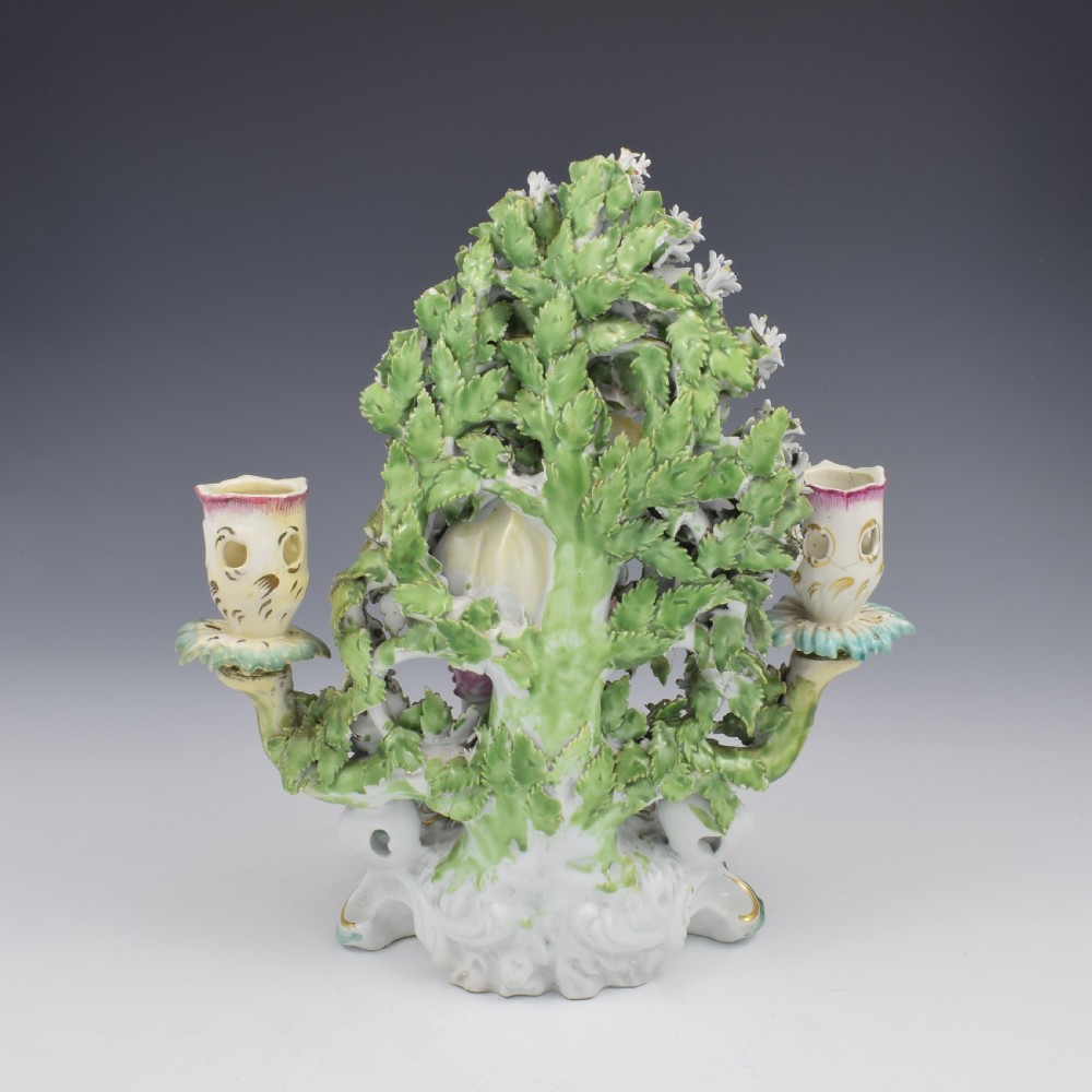 A pair of 18th-century Derby porcelain candlestick figures depicting children standing before flowering bocage trees set on scrollwork bases. View IX