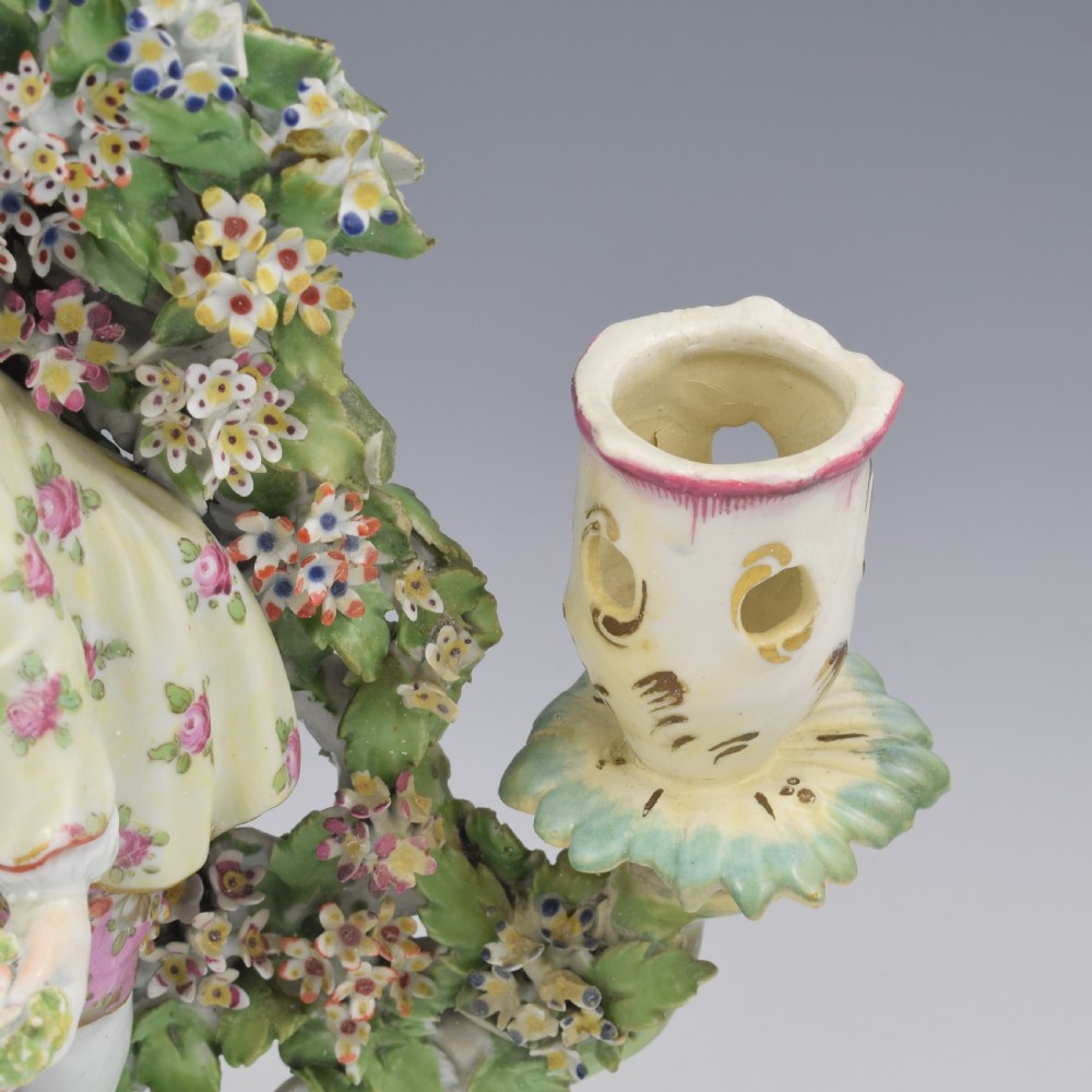 A pair of 18th-century Derby porcelain candlestick figures depicting children standing before flowering bocage trees set on scrollwork bases. View X
