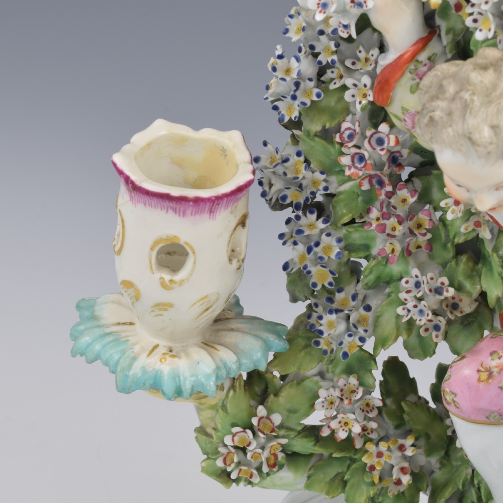 A pair of 18th-century Derby porcelain candlestick figures depicting children standing before flowering bocage trees set on scrollwork bases. View XI
