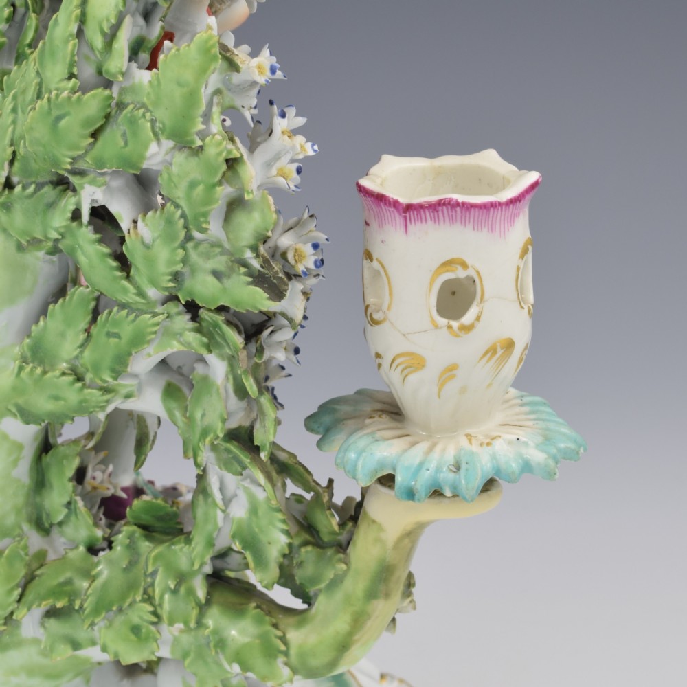 A pair of 18th-century Derby porcelain candlestick figures depicting children standing before flowering bocage trees set on scrollwork bases. View XII