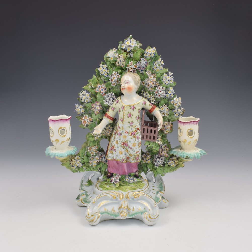 A pair of 18th-century Derby porcelain candlestick figures depicting children standing before flowering bocage trees set on scrollwork bases. View XIV