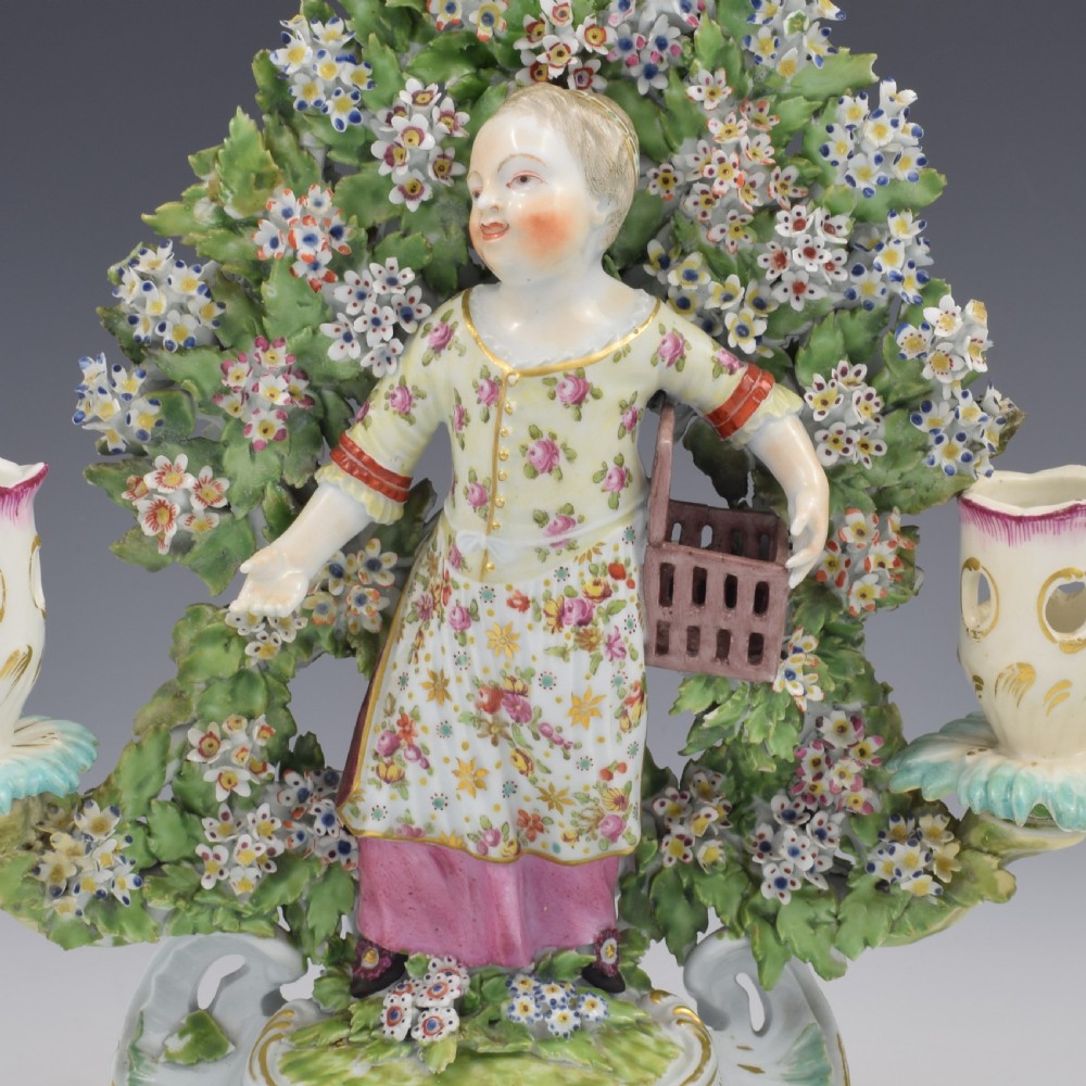 A pair of 18th-century Derby porcelain candlestick figures depicting children standing before flowering bocage trees set on scrollwork bases. View XV
