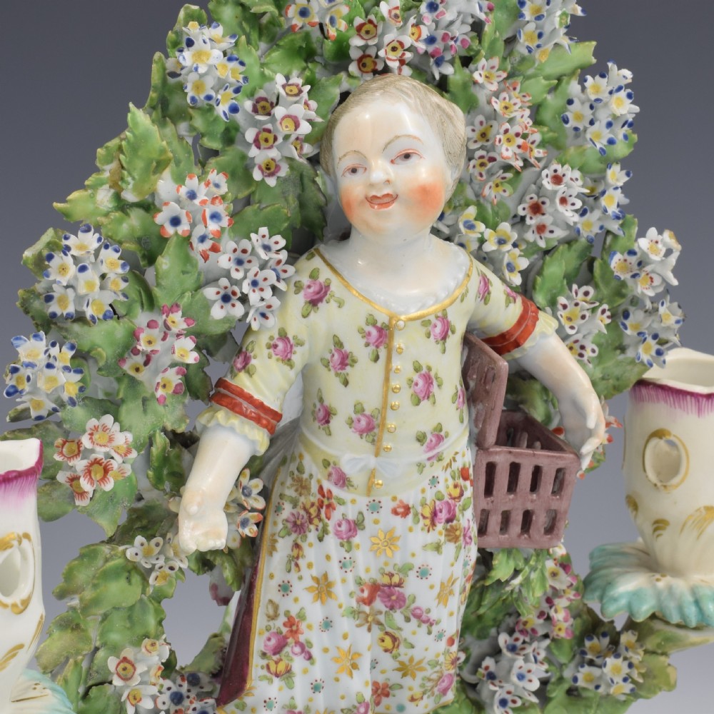 A pair of 18th-century Derby porcelain candlestick figures depicting children standing before flowering bocage trees set on scrollwork bases. View XVI