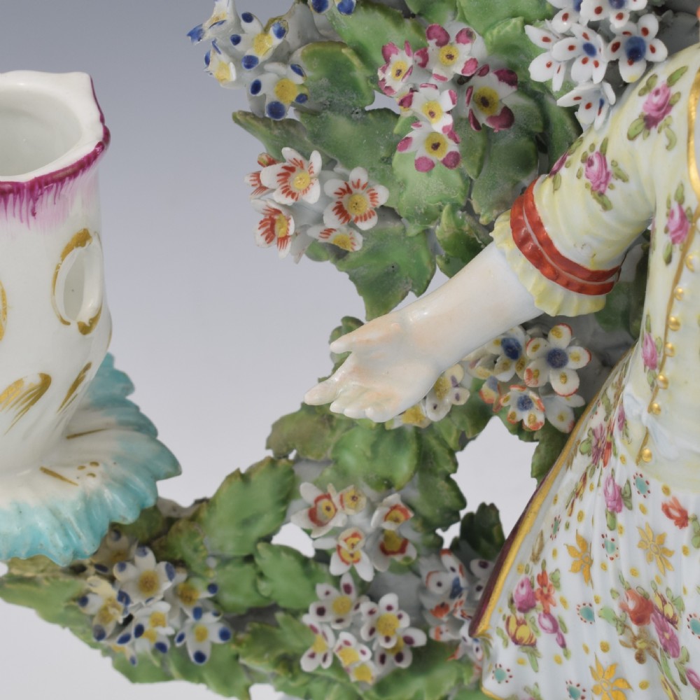 A pair of 18th-century Derby porcelain candlestick figures depicting children standing before flowering bocage trees set on scrollwork bases. View XVII
