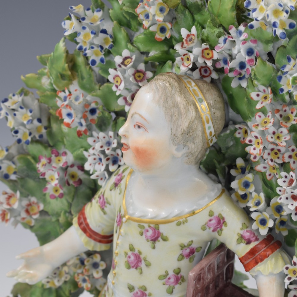 A pair of 18th-century Derby porcelain candlestick figures depicting children standing before flowering bocage trees set on scrollwork bases. View XVIII