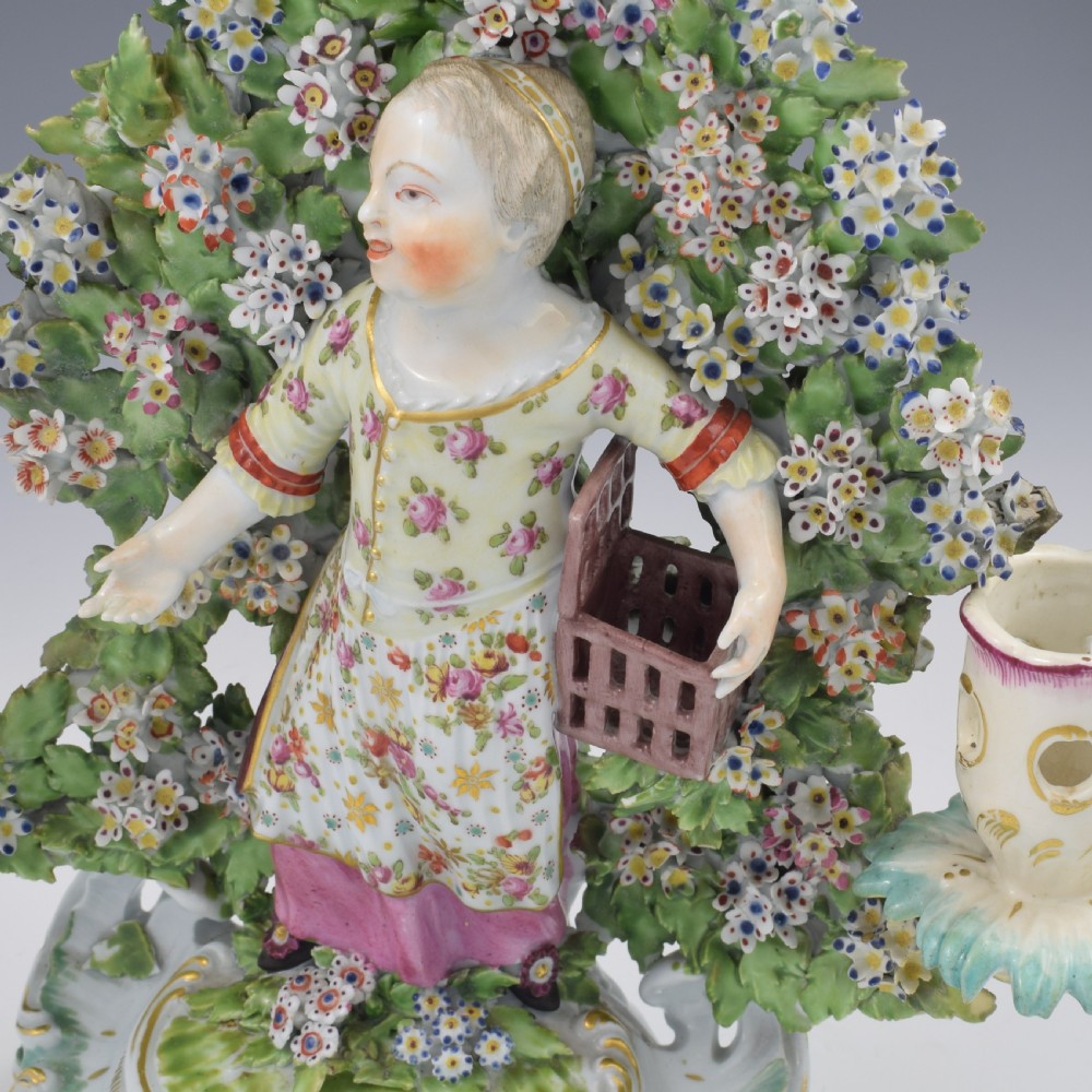A pair of 18th-century Derby porcelain candlestick figures depicting children standing before flowering bocage trees set on scrollwork bases. View XIX