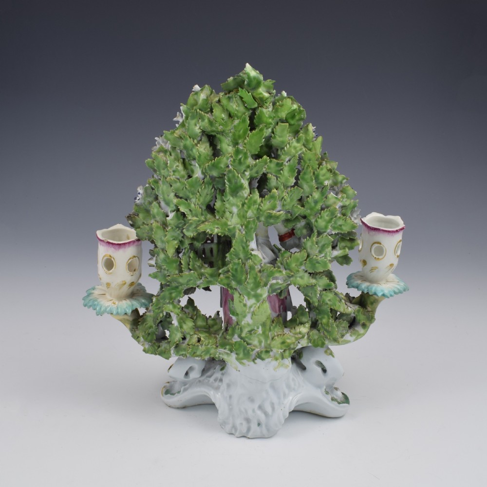 A pair of 18th-century Derby porcelain candlestick figures depicting children standing before flowering bocage trees set on scrollwork bases. View XX