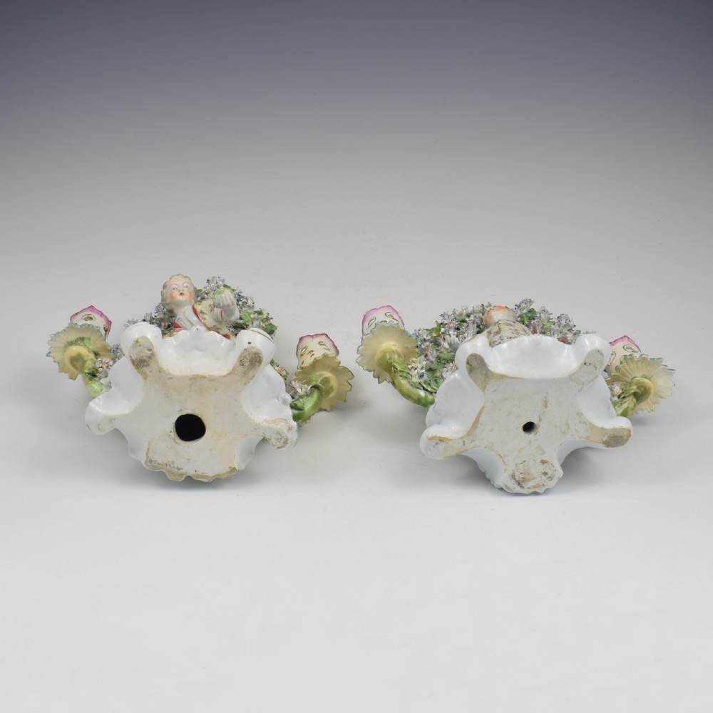 A pair of 18th-century Derby porcelain candlestick figures depicting children standing before flowering bocage trees set on scrollwork bases. View XXI