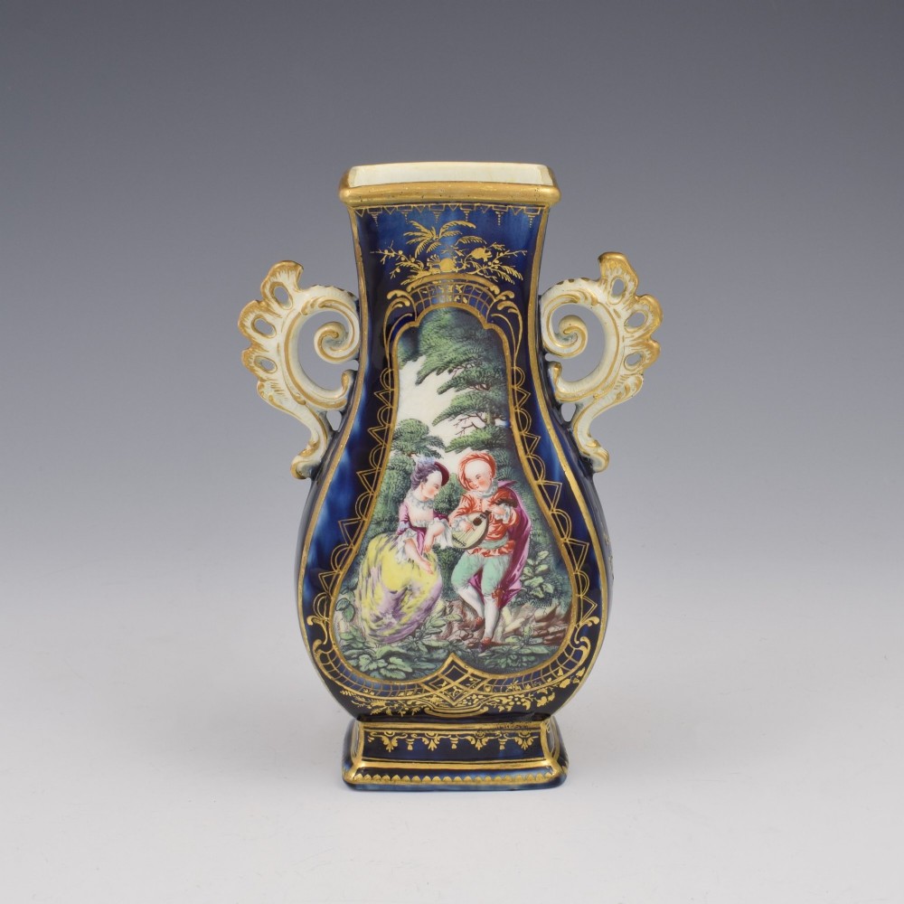 A cobalt blue soft-paste porcelain vase featuring elaborate gilt scroll handles, gilded leaf borders, and a central painted reserve depicting a gentleman playing a lute beside a lady in a landscape.