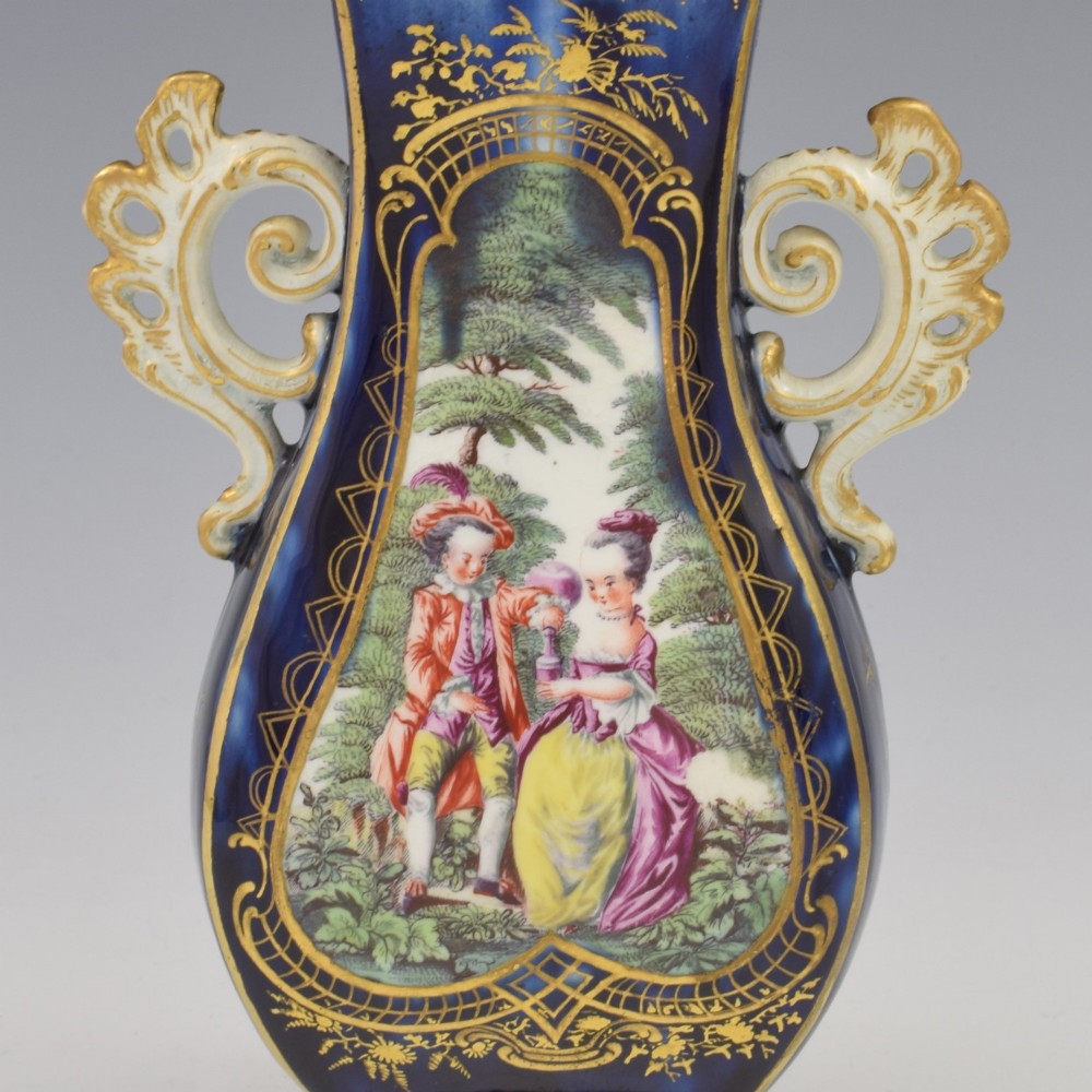 A cobalt blue soft-paste porcelain vase featuring elaborate gilt scroll handles, gilded leaf borders, and a central painted reserve depicting a gentleman playing a lute beside a lady in a landscape. View X