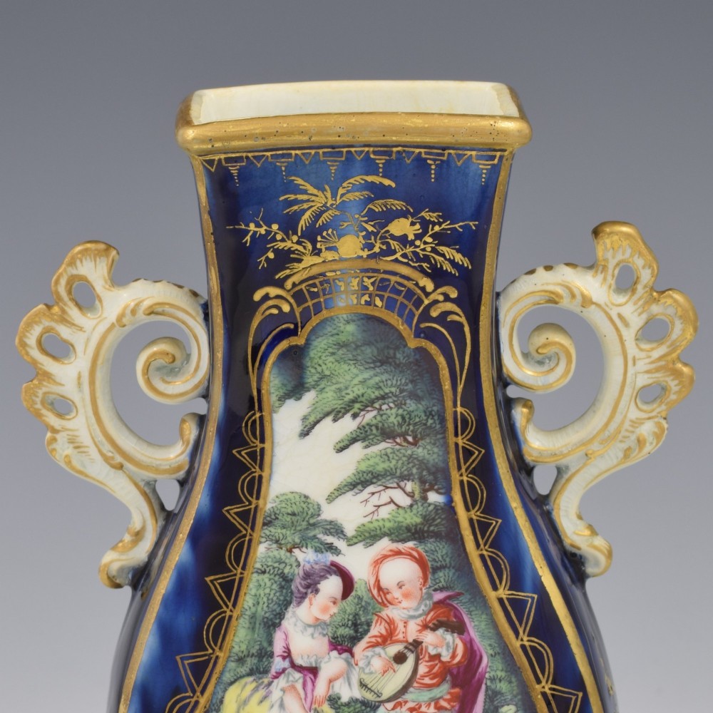 A cobalt blue soft-paste porcelain vase featuring elaborate gilt scroll handles, gilded leaf borders, and a central painted reserve depicting a gentleman playing a lute beside a lady in a landscape. View XI
