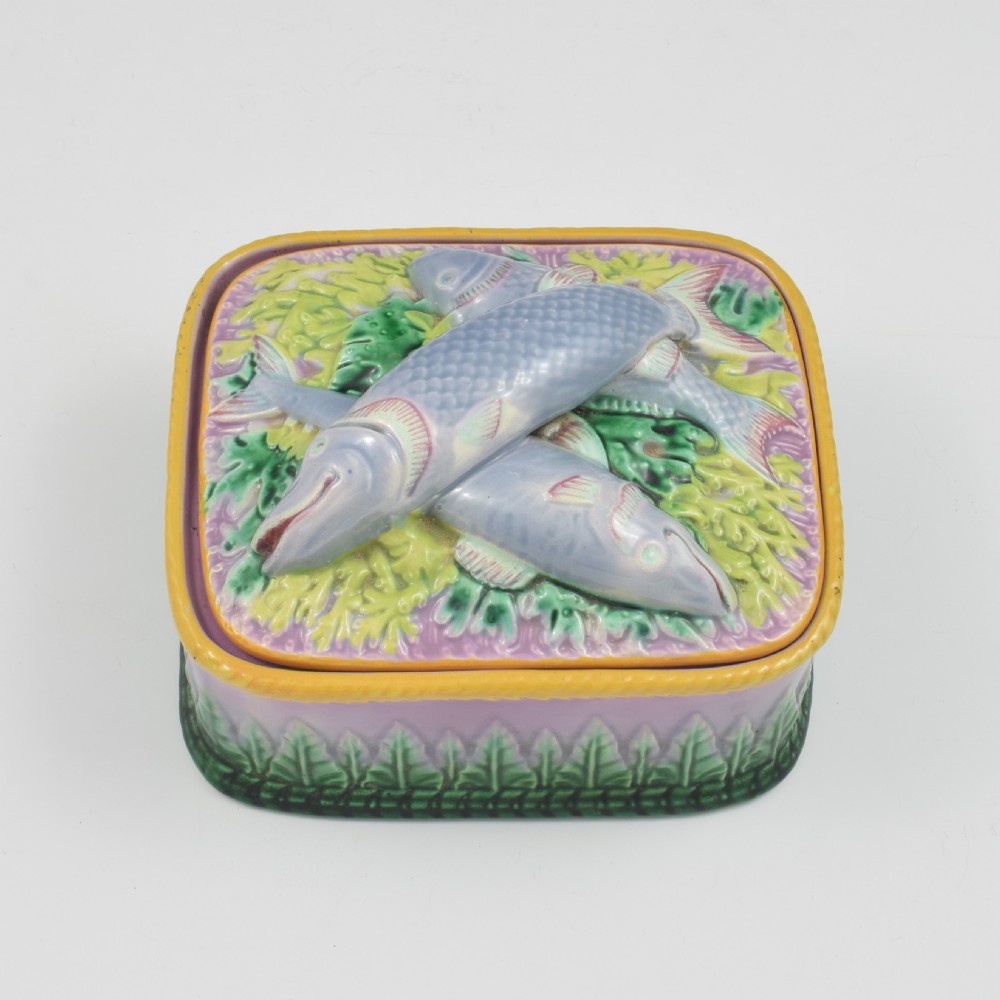 A rectangular Victorian majolica lidded box in lilac and pink featuring relief-moulded fish resting on seaweed with a yellow rim and green leaf base.