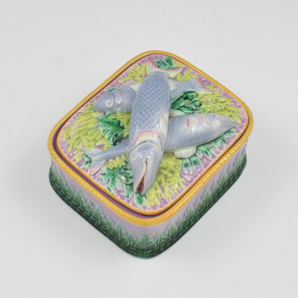 A rectangular Victorian majolica lidded box in lilac and pink featuring relief-moulded fish resting on seaweed with a yellow rim and green leaf base. View II