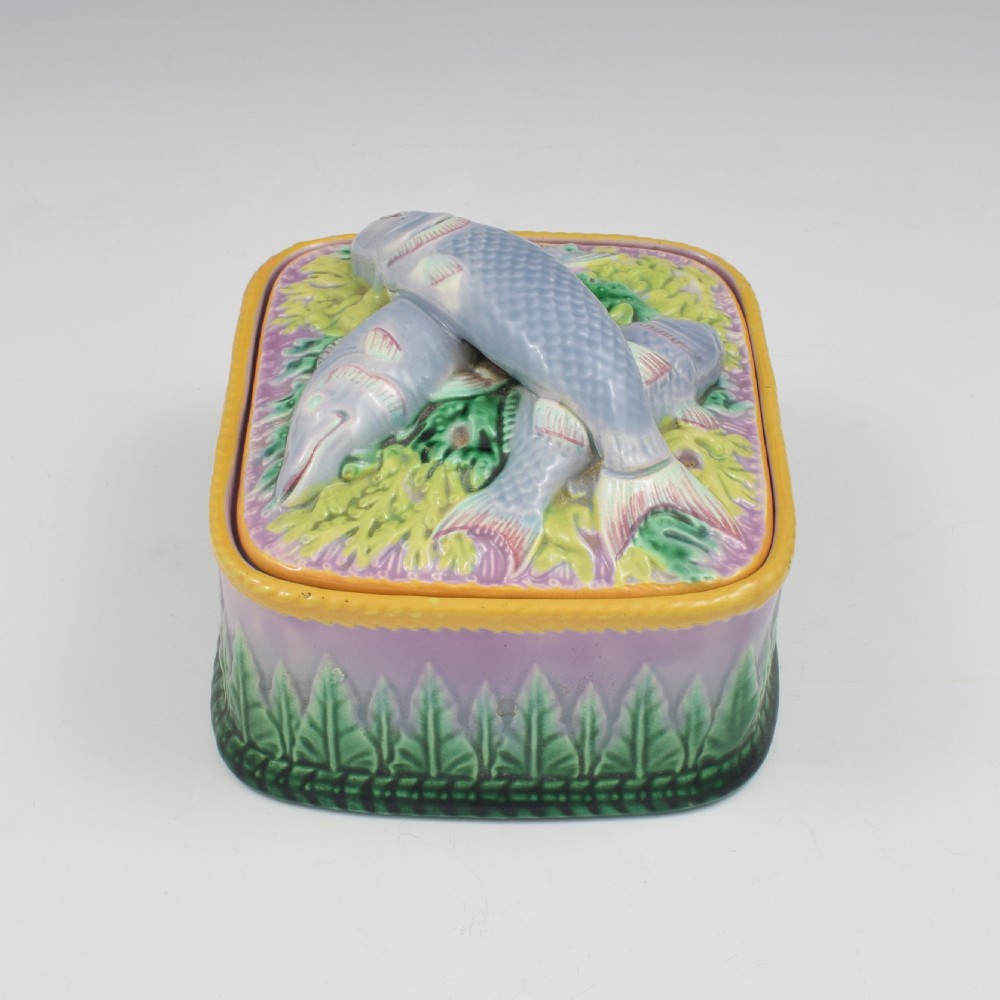A rectangular Victorian majolica lidded box in lilac and pink featuring relief-moulded fish resting on seaweed with a yellow rim and green leaf base. View V