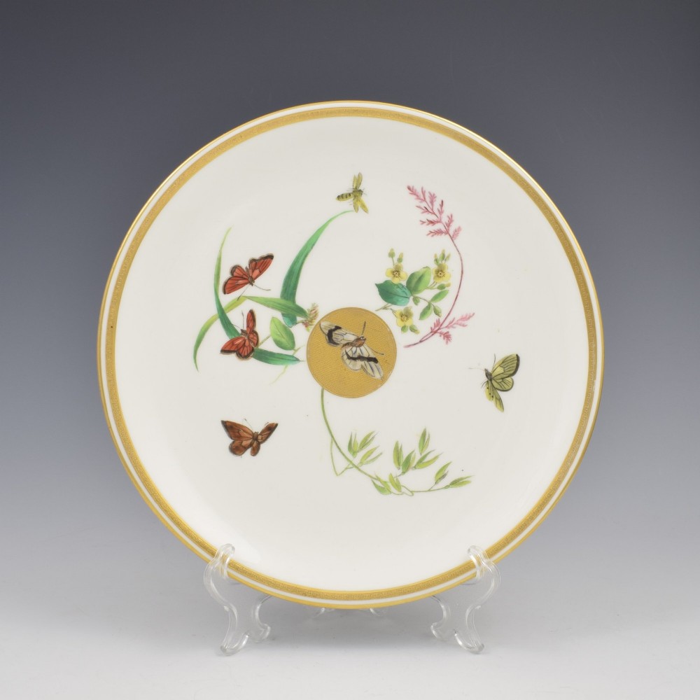 A circular cream porcelain dessert plate decorated with a central gilded sun motif, hand-painted butterflies, botanical sprigs, and a gilded rim.