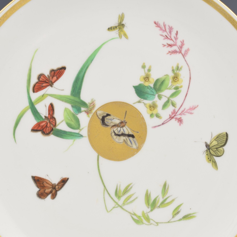 A circular cream porcelain dessert plate decorated with a central gilded sun motif, hand-painted butterflies, botanical sprigs, and a gilded rim. View II