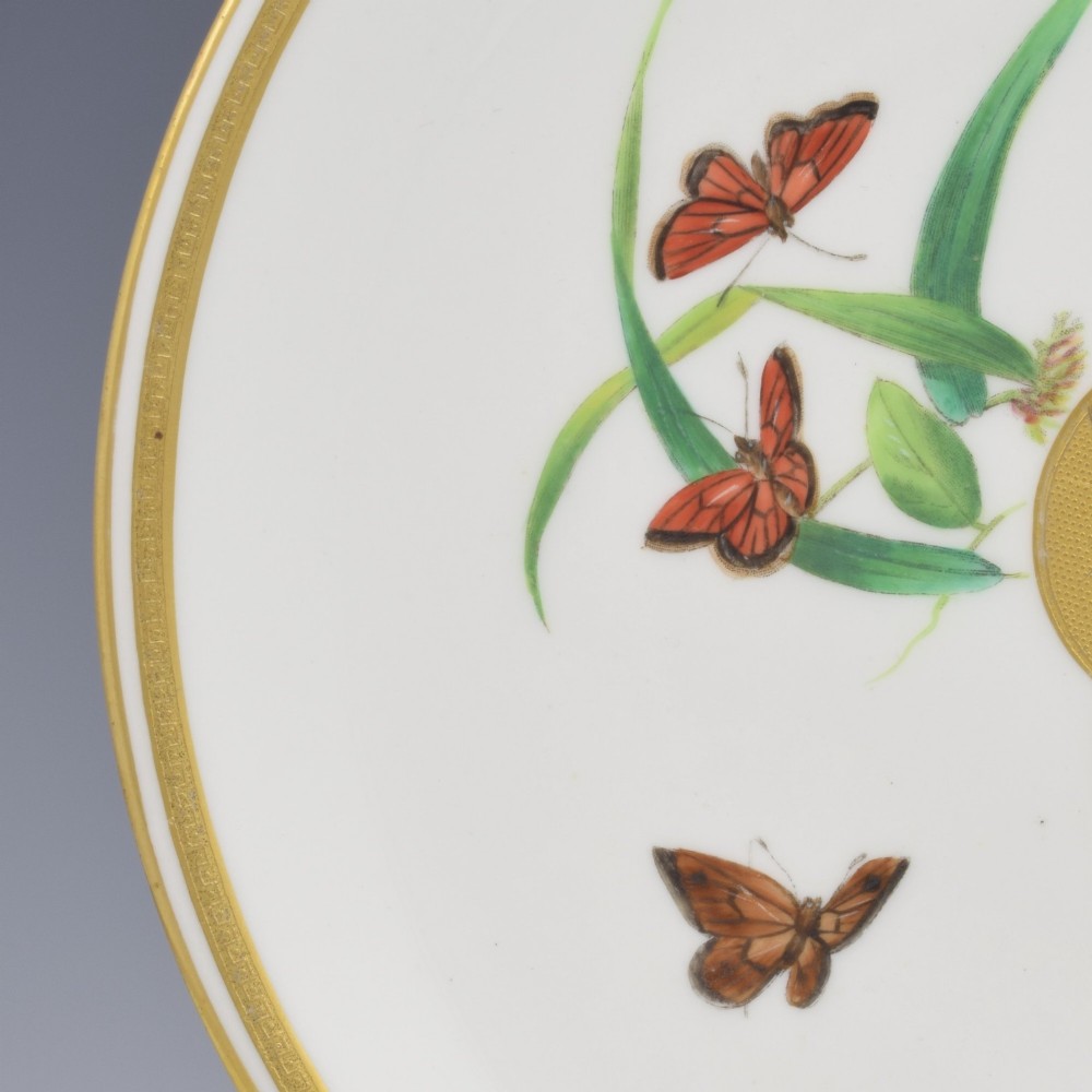 A circular cream porcelain dessert plate decorated with a central gilded sun motif, hand-painted butterflies, botanical sprigs, and a gilded rim. View IV