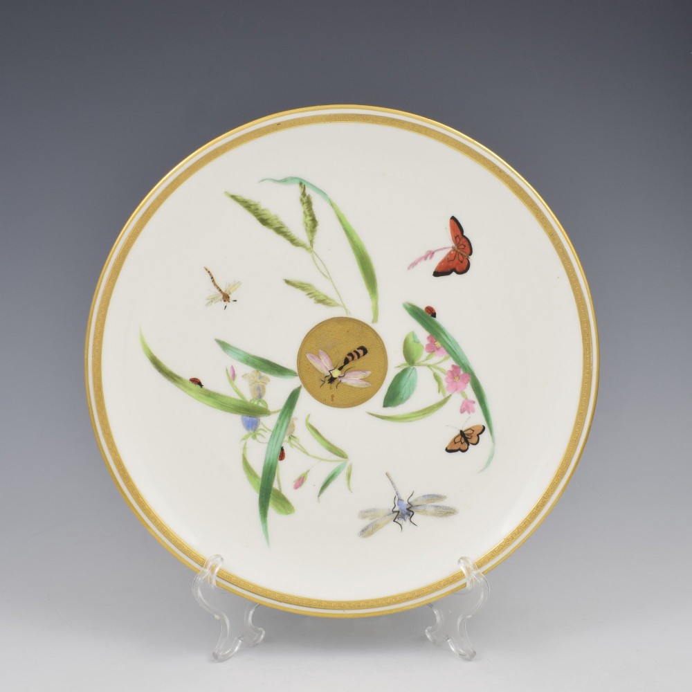 A white porcelain dessert plate decorated with hand-painted insects, green foliage, pink flowers, and a central gilt sun medallion with a gilded rim.