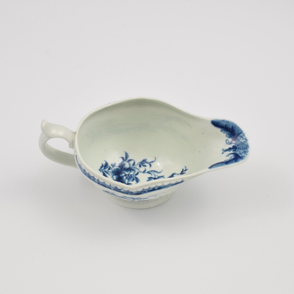 An eighteenth-century blue and white soft-paste porcelain sauce boat with a moulded fluted body, scroll handle, and underglaze blue landscape decoration. View X