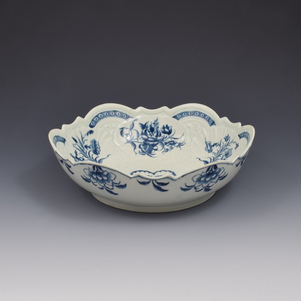 An eighteenth-century circular soft-paste porcelain junket dish featuring a scalloped rim, relief shell moulding, and painted blue floral motifs. View V