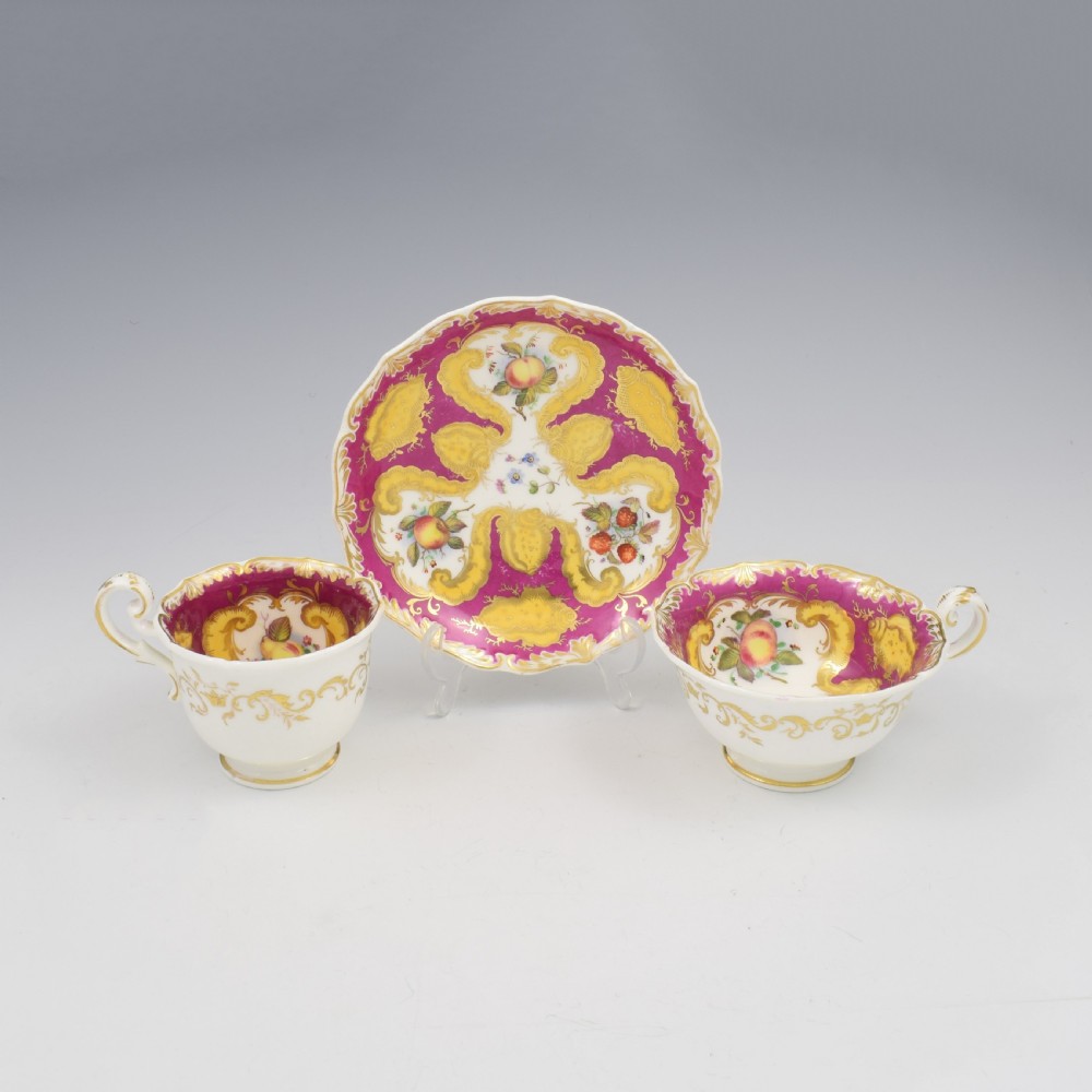 An antique English porcelain tea trio comprising a teacup, coffee cup, and saucer decorated with magenta ground, gilded scrollwork, and hand-painted fruit panels.