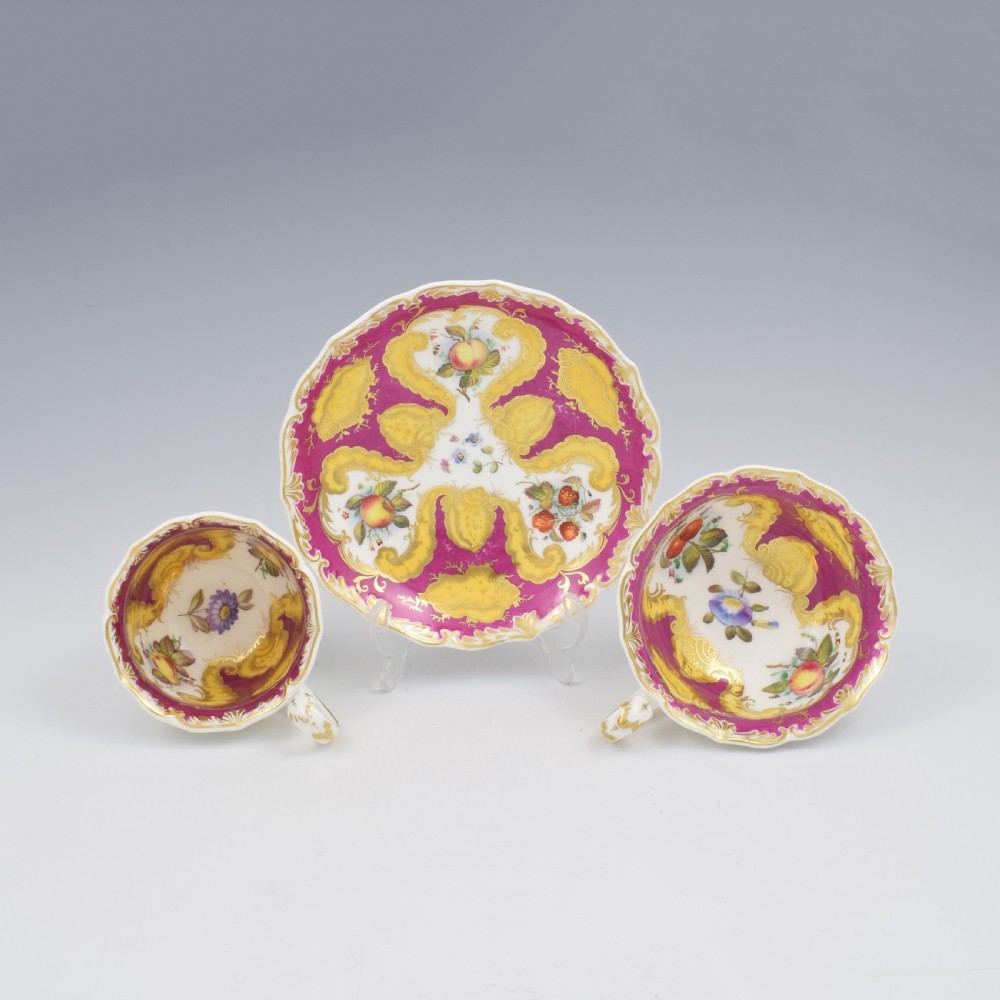 An antique English porcelain tea trio comprising a teacup, coffee cup, and saucer decorated with magenta ground, gilded scrollwork, and hand-painted fruit panels. View II