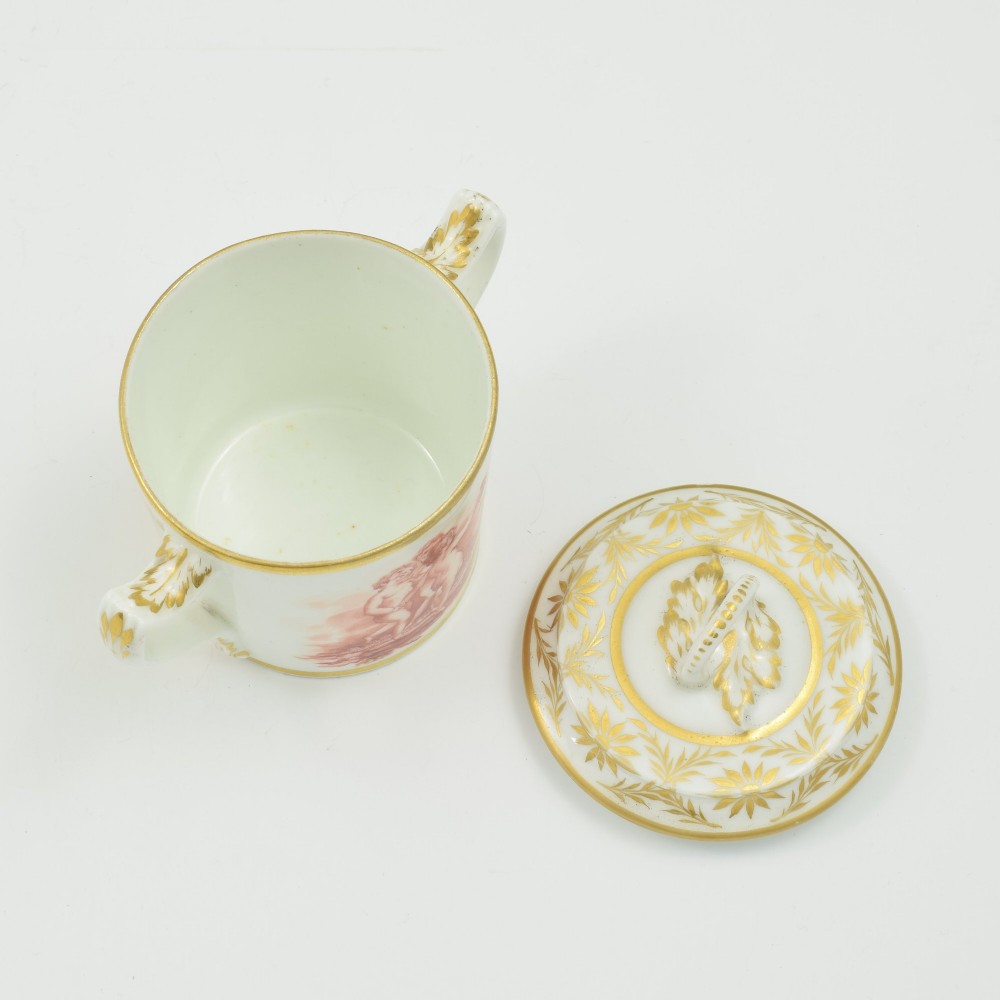 A white Regency period Bloor Derby porcelain chocolate cup featuring twin handles, a fitted lid with a loop finial, hand-painted sepia putti, and gold gilded leaf borders. View X