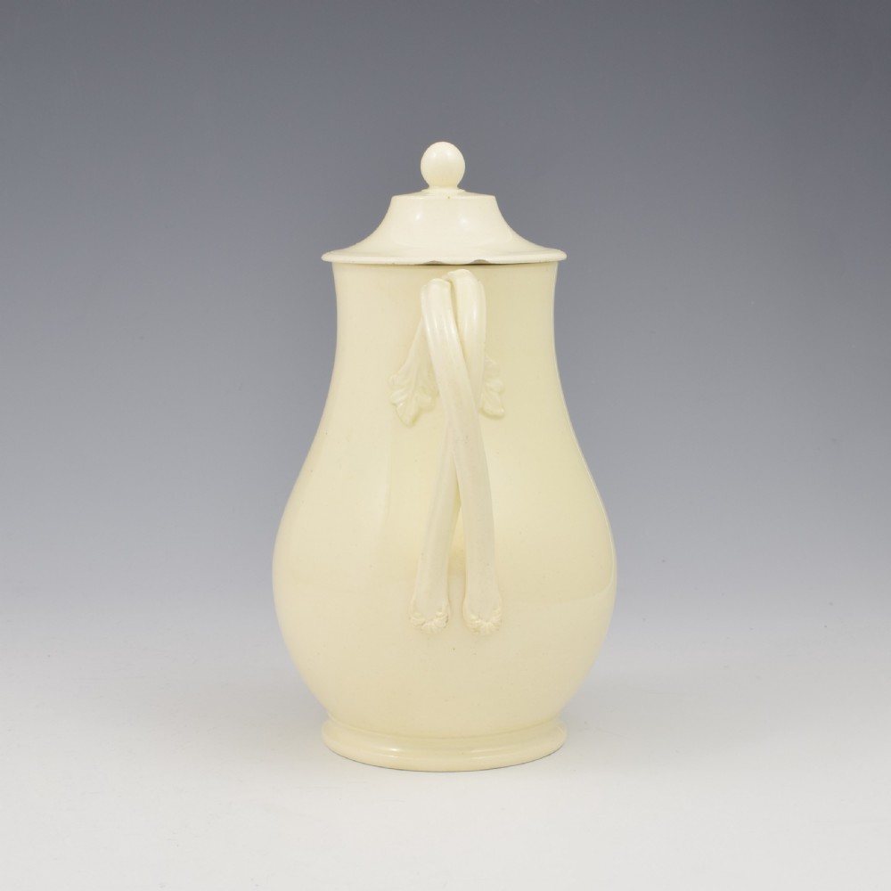 An 18th-century creamware hot water jug featuring a baluster-shaped earthenware body, a fitted dome cover with a ball finial, and a loop handle. View V