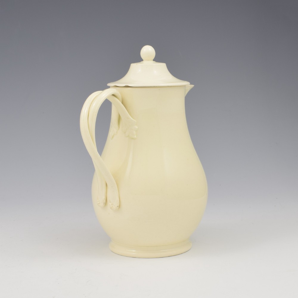 An 18th-century creamware hot water jug featuring a baluster-shaped earthenware body, a fitted dome cover with a ball finial, and a loop handle. View VI